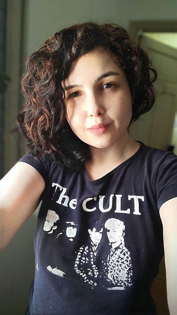 Selfie of a white-skinned woman with shoulder-length curly dark hair and brown eyes.